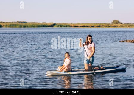 Young daughter and mother sitting together on sup board rowing with paddle on little lake with green reeds and trees in background. Active holidays Stock Photo