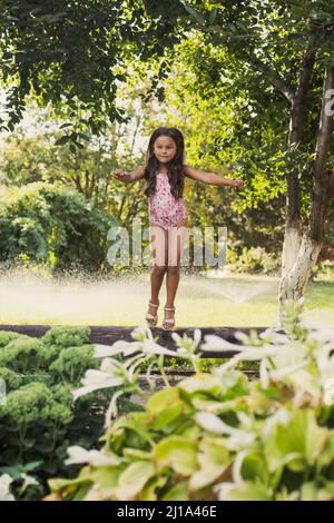 Happy young Caucasian girl balancing on log looking at camera surprisingly in backyard with green trees and water sprinklers in background in daytime