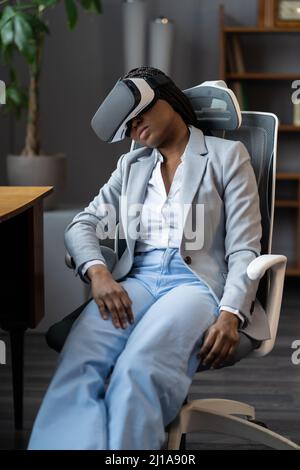 Cybersickness. Tired african american female employee wearing VR headset sleeping in office chair Stock Photo