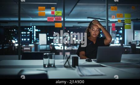 Overworked African American Businesswoman Working on Laptop Computer in Big City Office Late in the Evening. Tired Stressed Female Entrepreneur trying Stock Photo