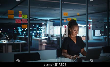 Overworked African American Businesswoman Working on Laptop Computer in Big City Office Late in the Evening. Tired Stressed Female Entrepreneur trying Stock Photo