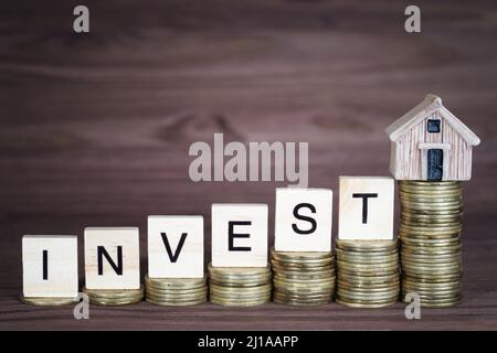 Word INVEST made from wooden letter blocks and a miniature house placed on ascending coins stacks. Creative home investment concept. Stock Photo