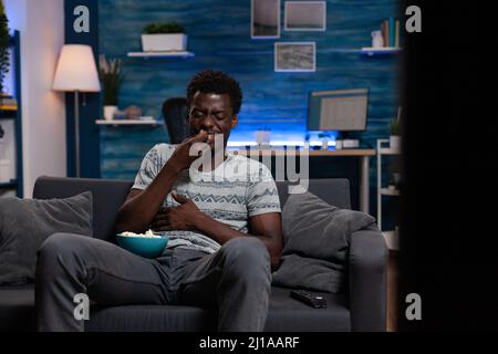 Smiling young person resting on couch holding bowl with popcorn while watching comedy movie on television relaxing afer job at home in living room. Guy enjoying fun activity during free time Stock Photo