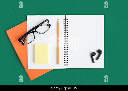 Notebook with pencil, glasses, wireless headphones. Open notebook on dark green color background, spiral notepad. Office desk. Copy Space Stock Photo