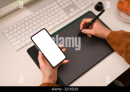 Top view, Female graphic designer's hand drawing on graphic tablet with stylus pen and holding smartphone white screen mockup over modern office desk. Stock Photo