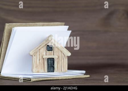 Close-up of a tiny house model placed on a small open book with blank pages against a wooden table background. Making a household manual. Stock Photo