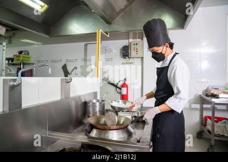 Asian male chef cook wearing face protective medical mask for protection from virus disease cooking food in frying pan at restaurant kitchen. Stock Photo