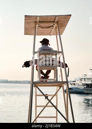 Lifeguard is sitting on wooden chair on tower watching the sea in Dubai. Boat is moored, the sea is calm. Nobody is in water Stock Photo