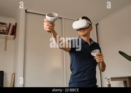 Young man in casual clothes plays virtual games at home. Man in virtual reality VR helmet compensates lack of movement. Stock Photo