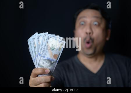 Asian handsome man holding bunch of dollars banknotes on black background scared and amazed with open mouth for surprise, disbelief face Stock Photo