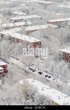 Snow-covered roofs of apartment buildings after snowfall the day before. Snow road between houses. Fluffy snow lies on branches of trees. Cars are Stock Photo