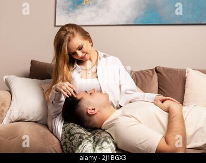 Happy couple relaxing on couch. Handsome man lying on sofa and putting head on girlfriend or wife knees. Beautiful woman holding man hand and smiling. Tenderness, love concept. High quality photo Stock Photo