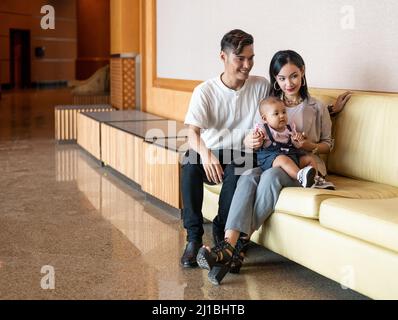 Malay Malaysian Muslim Young Couple with one year old child daughter seated on a long light brown settee chair Stock Photo