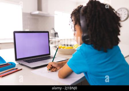 Hispanic boy wearing headphones while writing and attending online class through laptop Stock Photo