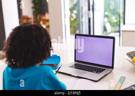 Rear view of hispanic boy attending online class through laptop while sitting at dining table Stock Photo