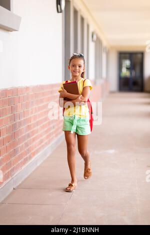 Full length portrait of caucasian elementary schoolgirl with books and backpack walking in corridor Stock Photo