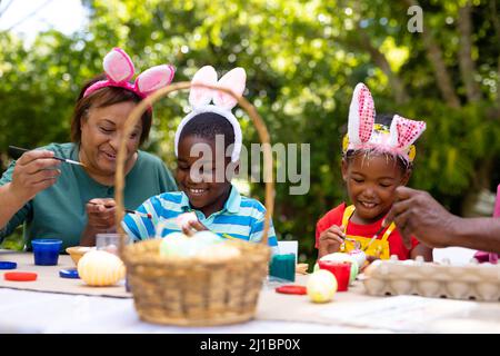 Smiling african american siblings and grandmother wearing bunny ears while painting eggs on easter Stock Photo