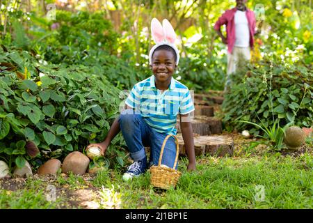 Portrait of smiling cute african american boy wearing bunny ears hiding easter egg in backyard Stock Photo