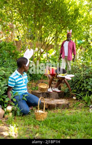 African american siblings in bunny ears hiding easter eggs with grandfather in backyard Stock Photo