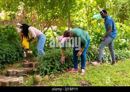 African american multi-generational family in bunny ears searching easter eggs in backyard Stock Photo