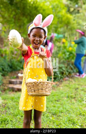Portrait of happy african american girl in bunny ears showing easter egg while family in backyard Stock Photo