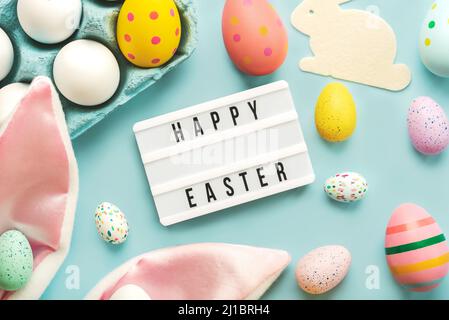 Happy Easter. Light box with the text Happy Easter,colored easter painted eggs and rabbit ears over blue background Stock Photo
