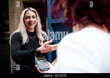 Alternative woman using wireless terminal for contactless payment with smart watch at a coffee shop. Stock Photo