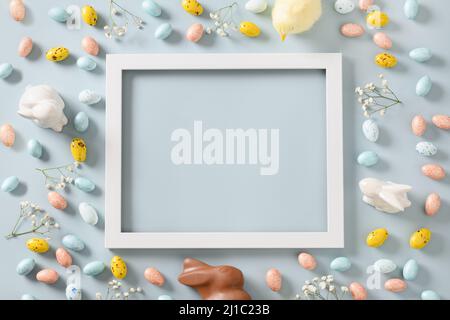Easter frame with sweet chocolate eggs and bunny, spring blossom flowers on blue background. Top view. Happy Easter greeting card with copy space. Stock Photo