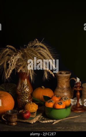 Vintage autumn still life with an earthenware jug with pumpkins, persimmons and wheat grain ears on an old wooden surface on a black background with s
