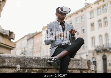 Successful african businessman in stylish wear using VR headset while sitting on city street. Young male person gesturing with hands in air while touching virtual screen. Stock Photo