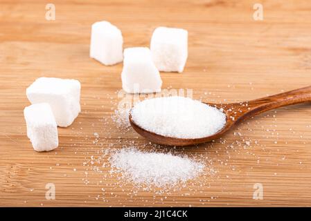 Lump sugar and a wooden spoon with white sugar on a wooden background. Sugar is scattered on a wooden surface. The sugar in the background is blurry. Stock Photo