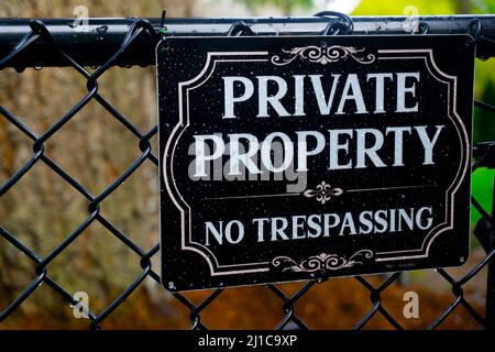 A sign reading Private Property No Tresspassing sits on a chain link fence in front of a natural background of trees and grass. Stock Photo