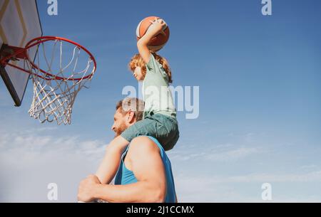 Father and son playing basketball. Sports Concept. Boy child sitting on the dad shoulders, throwing basketball ball into basket, side view on sky Stock Photo