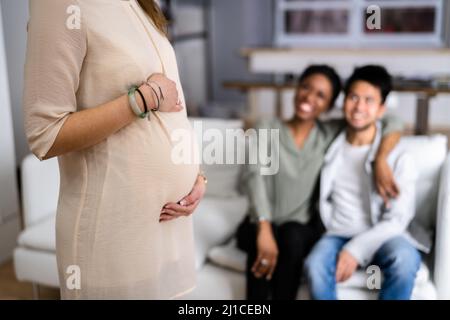 Pregnant Woman Touching Her Belly's Woman Standing In Front Of Smiling Young Couple Sitting Stock Photo