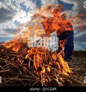 Firefighter ecologist fighting fire in field with cloudy sky on background. Low angle view of male environmentalist holding bucket and pouring water on burning dry grass. Natural disaster concept. Stock Photo