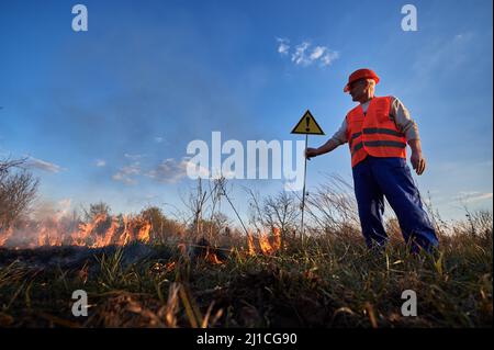 Fireman ecologist extinguishing fire in field in evening. Man in orange vest and helmet near burning grass with smoke, holding warning sign with exclamation mark. Natural disaster concept. Stock Photo