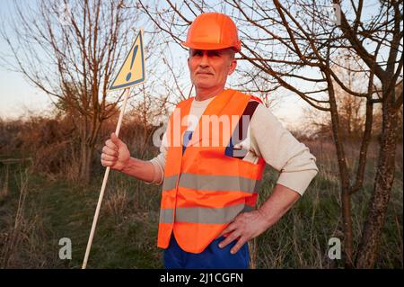 Fireman ecologist extinguishing fire in field in evening. Man in orange work vest and safety helmet near burning grass with smoke, holding warning sign with exclamation mark. Natural disaster concept. Stock Photo