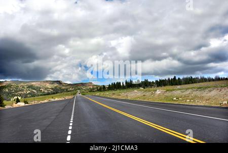 Blacktop highway disappears into the mountains of Beartooth Pass Scenic Byway.  Stormy clouds threaten blue sky over mountains. Stock Photo