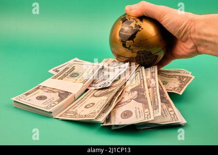 A pile of dollars and a man's hand holding a ball of the world with a green background. Concept money strangles the world. Photograph in horizontal fo Stock Photo