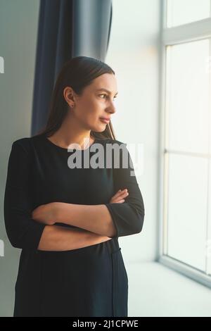 Young beautiful brunette girl or woman in black dress looks thoughtfully out window with her arms crossed over chest, vertical image Stock Photo