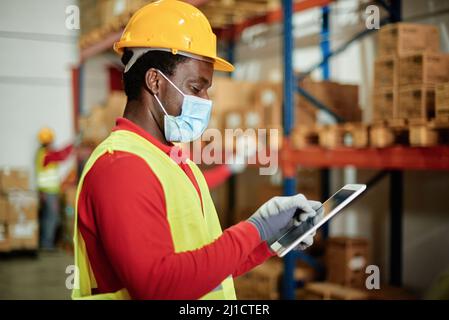 Afro male worker using a tablet checking the transport of a box in a warehouse wearing a safety mask and work waistcoat. Stock Photo