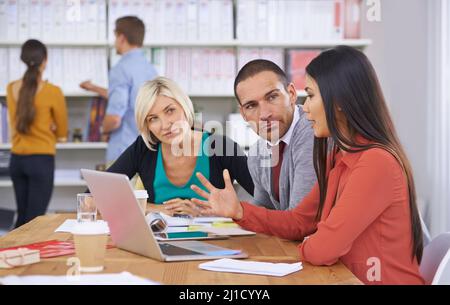 We have to come up with a solution. Three coworkers brainstorming around a laptop while their colleagues read files in the background. Stock Photo