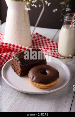 homemade chocolate donut and sponge cake covered with chocolate glaze and glass of milk on white rustic wooden surface Stock Photo