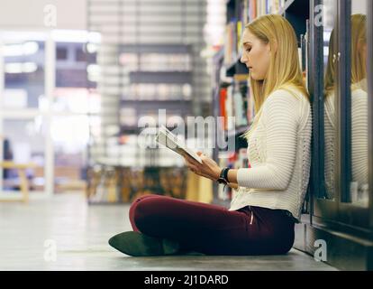 Shes diligent when it comes to her studies. Full length shot of an attractive young university reading a book while sitting on the library floor. Stock Photo