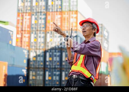 A senior elderly Asian worker engineer wearing safety vest and helmet standing and holding radio walkies talkie at logistic shipping cargo containers Stock Photo