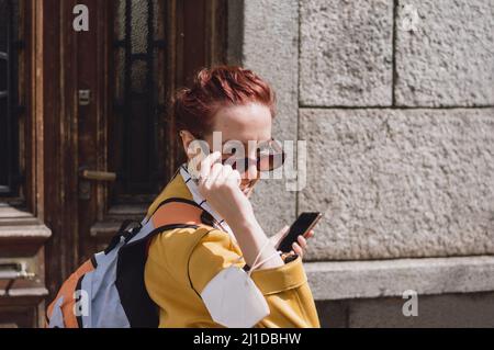 young caucasian woman walking in the street on a sunny day looking over her sunglasses with the phone in one hand, carrying a backpack and wearing a y Stock Photo