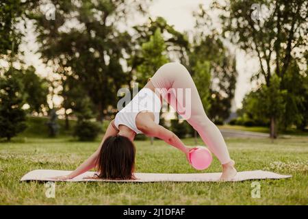 young fit woman pink sportswear and rubber rubber pilates ball does exercises and stands in down dog pose in park in summer Stock Photo