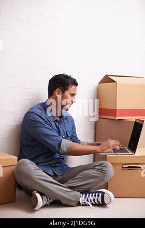 Hes moving house. A handsome young man using his laptop on the floor with boxes surrounding him. Stock Photo