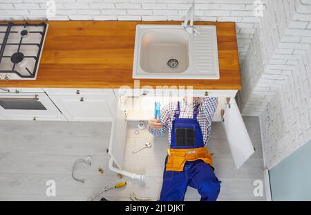 Plumber lying down on the floor under the kitchen sink drain and repairing pipes Stock Photo
