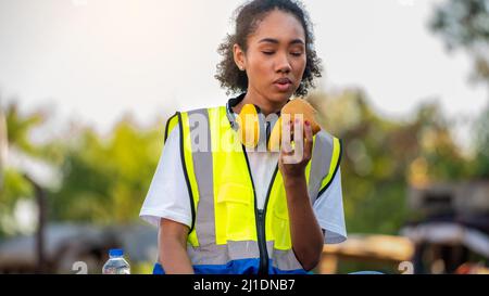 smile African american woman foreman worker  or woman maintenance engineer in reflective vest safety jacket sits down on old truck for relaxing, eats Stock Photo